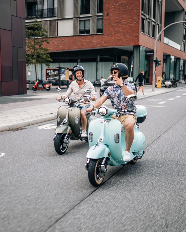 Zwei Freunde fahren mit Helm lachend auf zwei Elektrorollern über die Straße